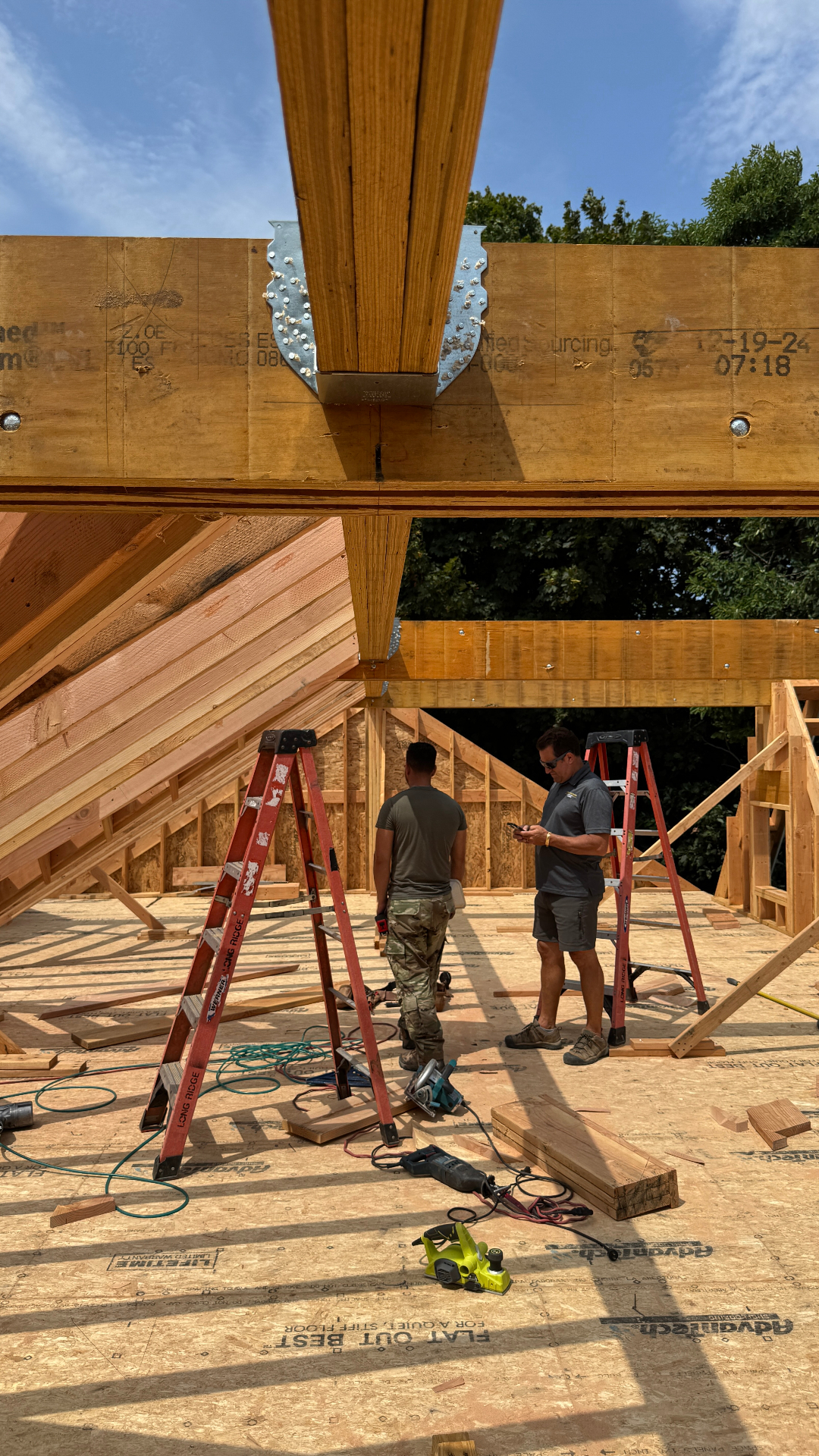 Two team members conferring beneath heavy timber ridge beams and steel connector plates during the framing stage of a new residence.