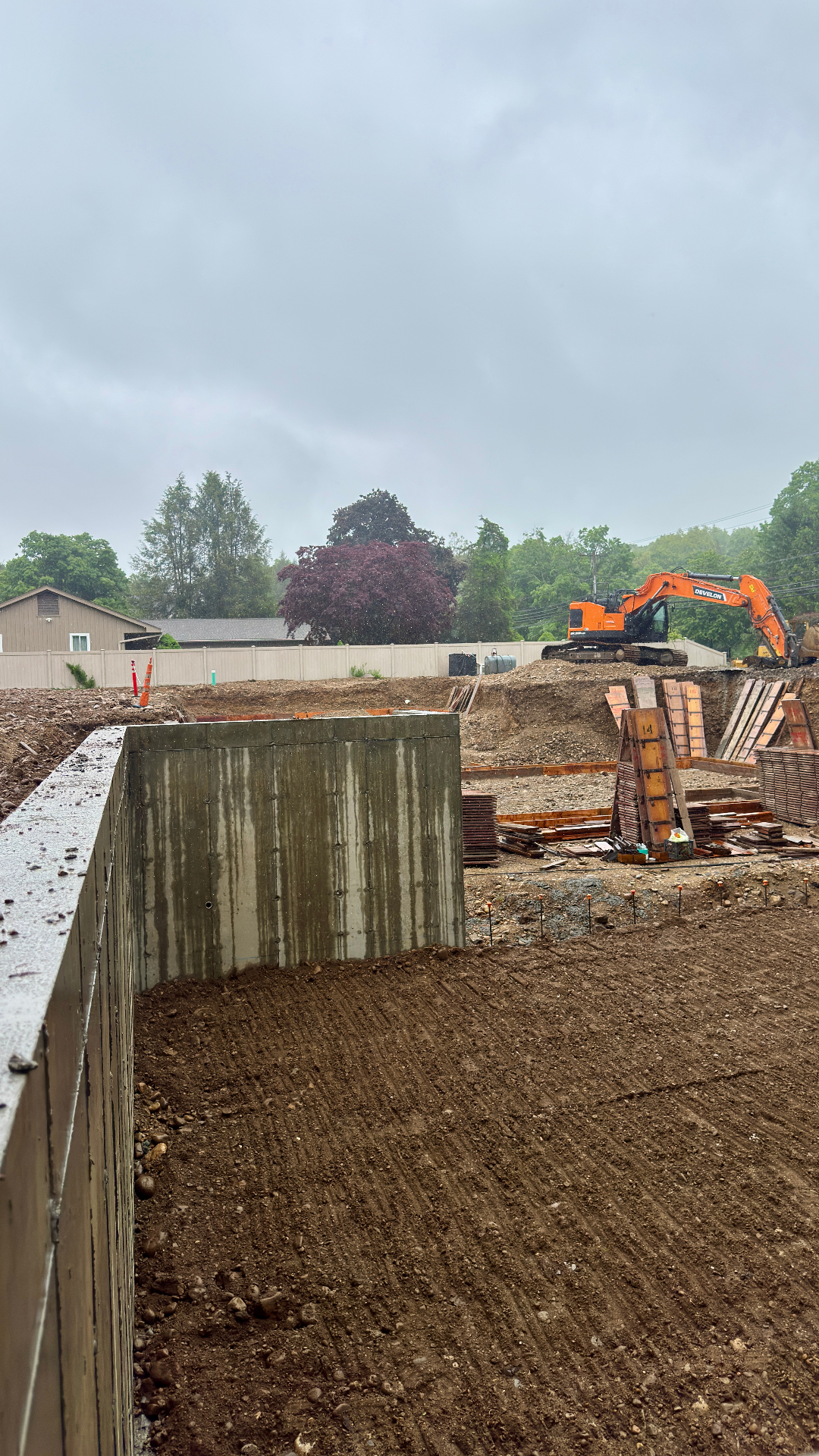 Completed poured-concrete foundation walls on a rainy day, with formwork panels stacked nearby and an excavator on the bank above.