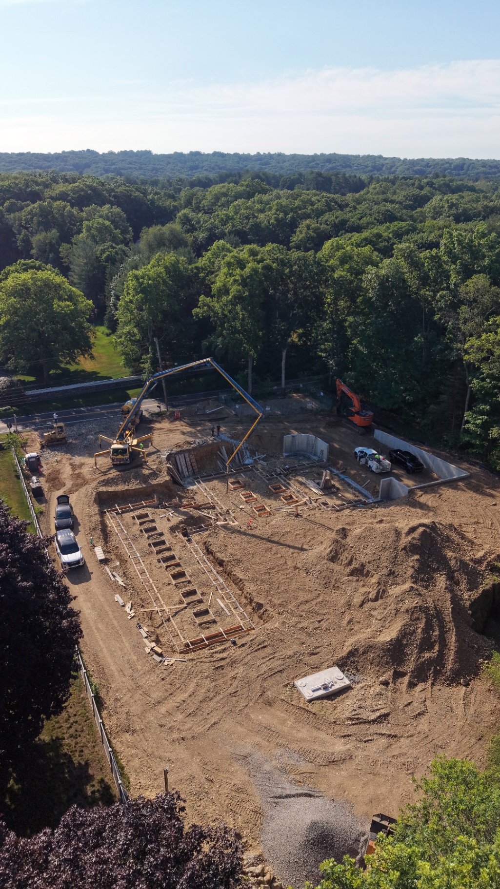 Aerial view of the foundation stage, with concrete walls, footing formwork, and a pump truck delivering concrete on a wooded lot.
