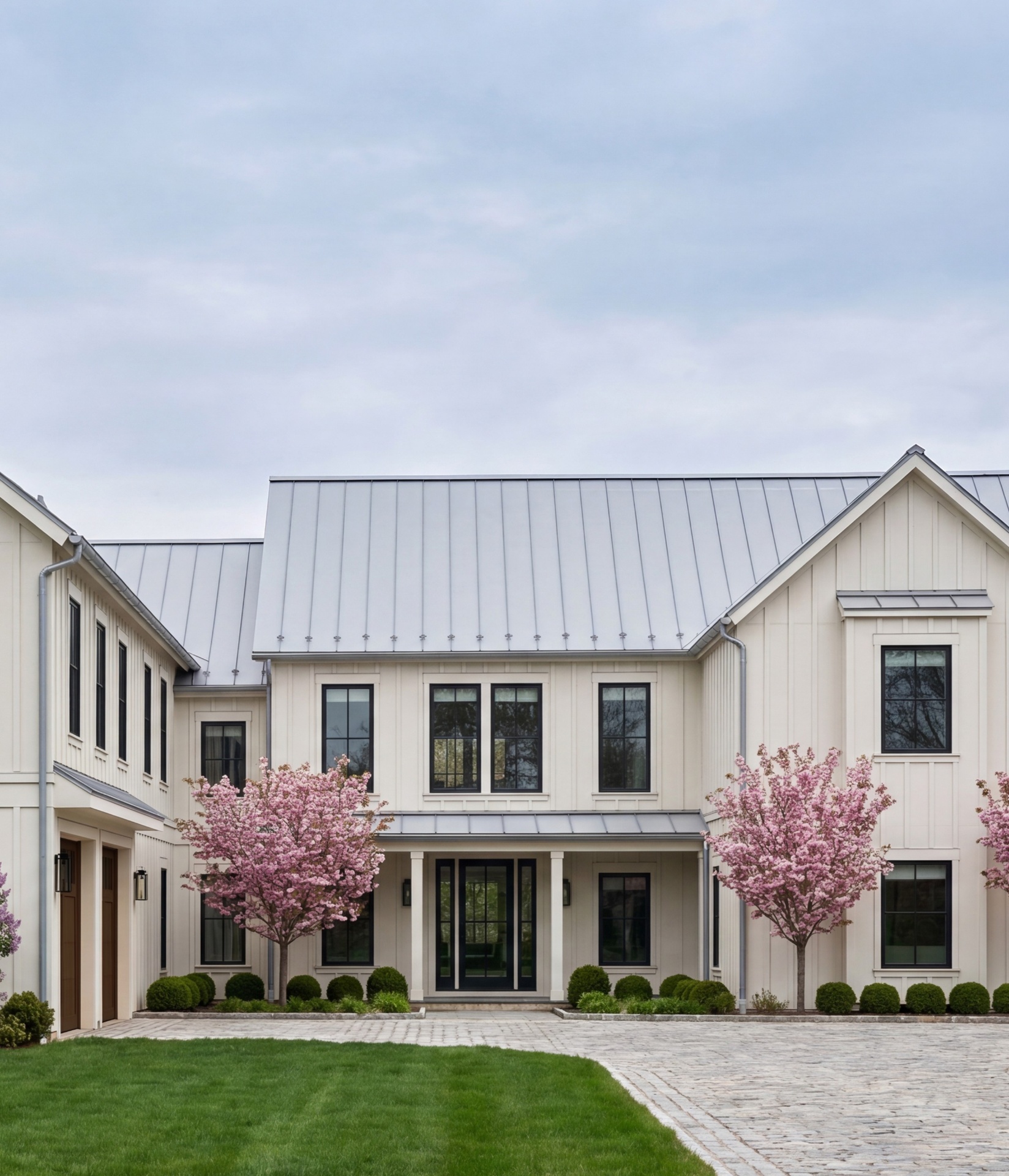 Completed board-and-batten residence with standing-seam metal roof, flowering cherry trees, and a cobblestone courtyard at handover.